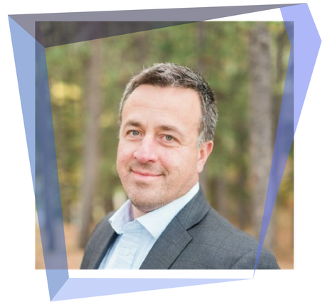 Man in suit smiling, framed in a blue geometric shape, with a blurred outdoor background.