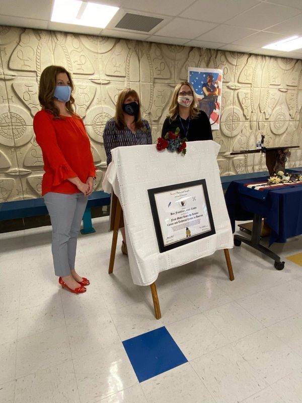 Three women wearing masks are standing around a table with a certificate on it.