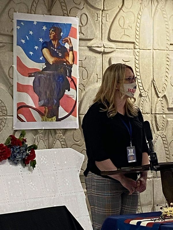 A woman wearing a mask is standing in front of a painting of a woman holding a microphone.