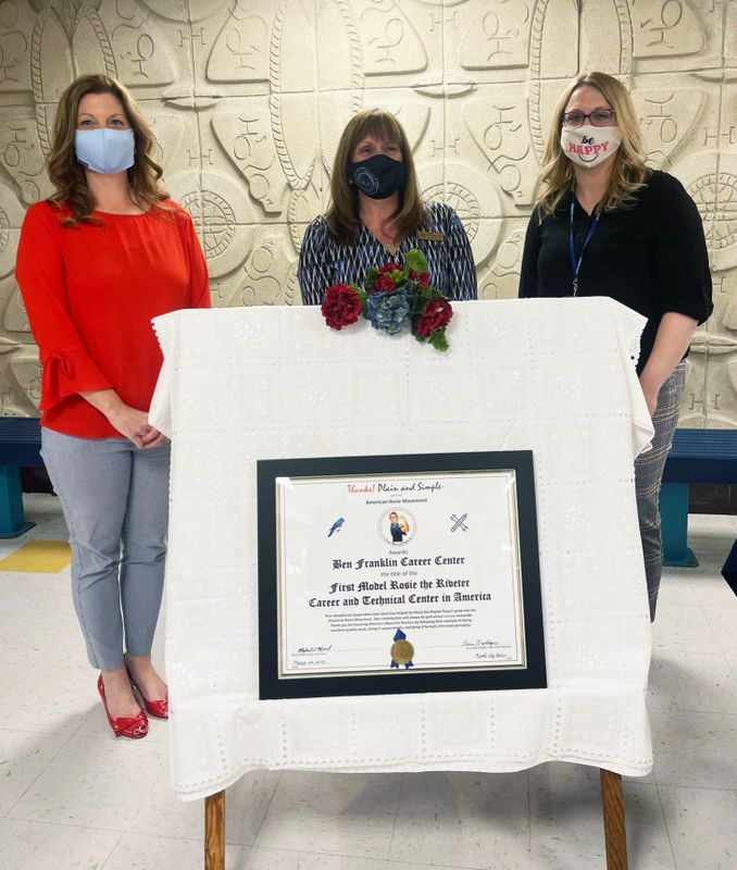 Three women wearing face masks are standing around a table with a certificate on it