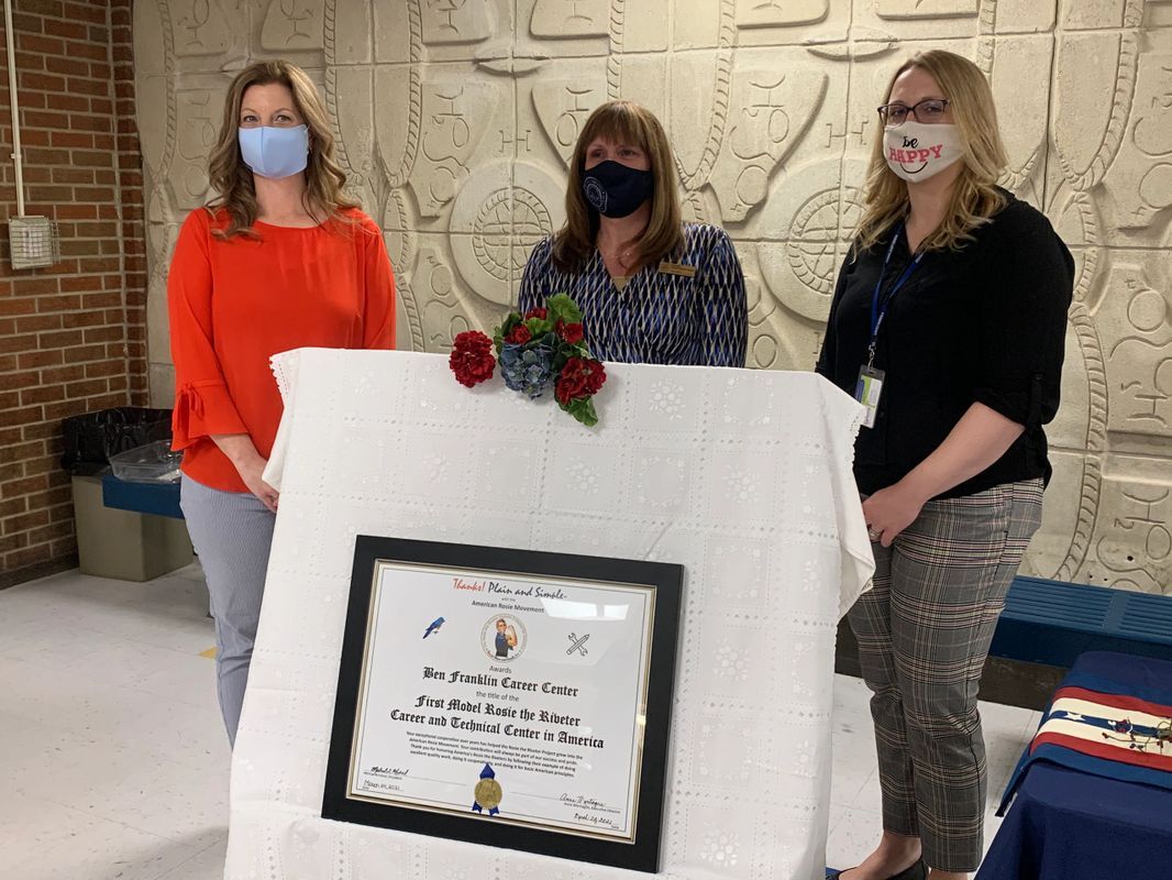 Three women wearing face masks are standing next to a plaque.