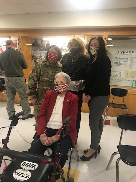 A group of people wearing face masks are standing around an elderly woman sitting on a walker.