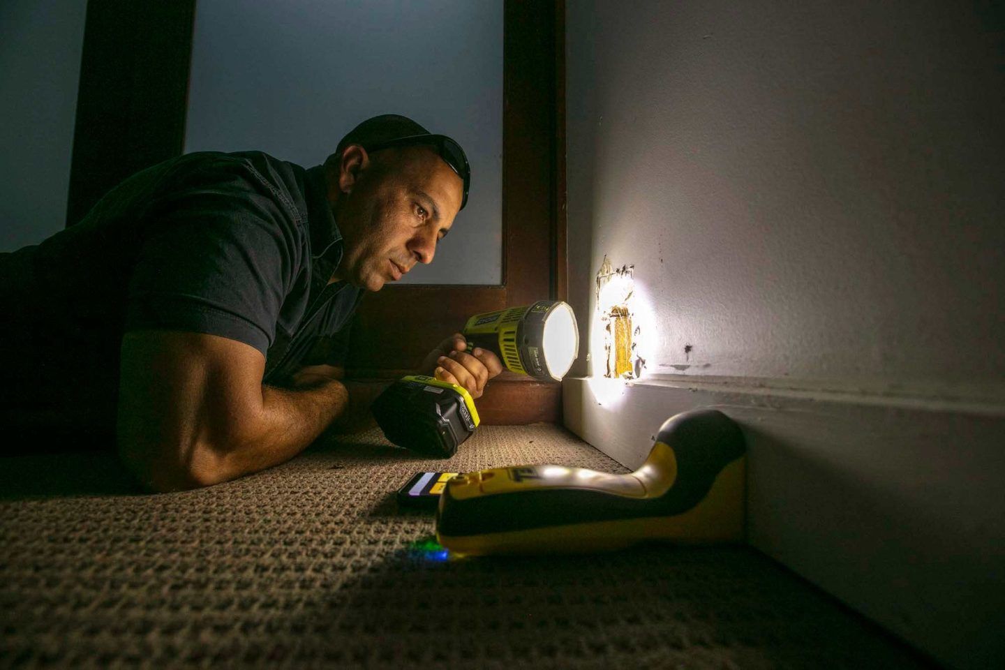 A Man is Laying on the Floor Looking at a Bug With a Flashlight — Superior Termite & Pest Control in Bundaberg, QLD