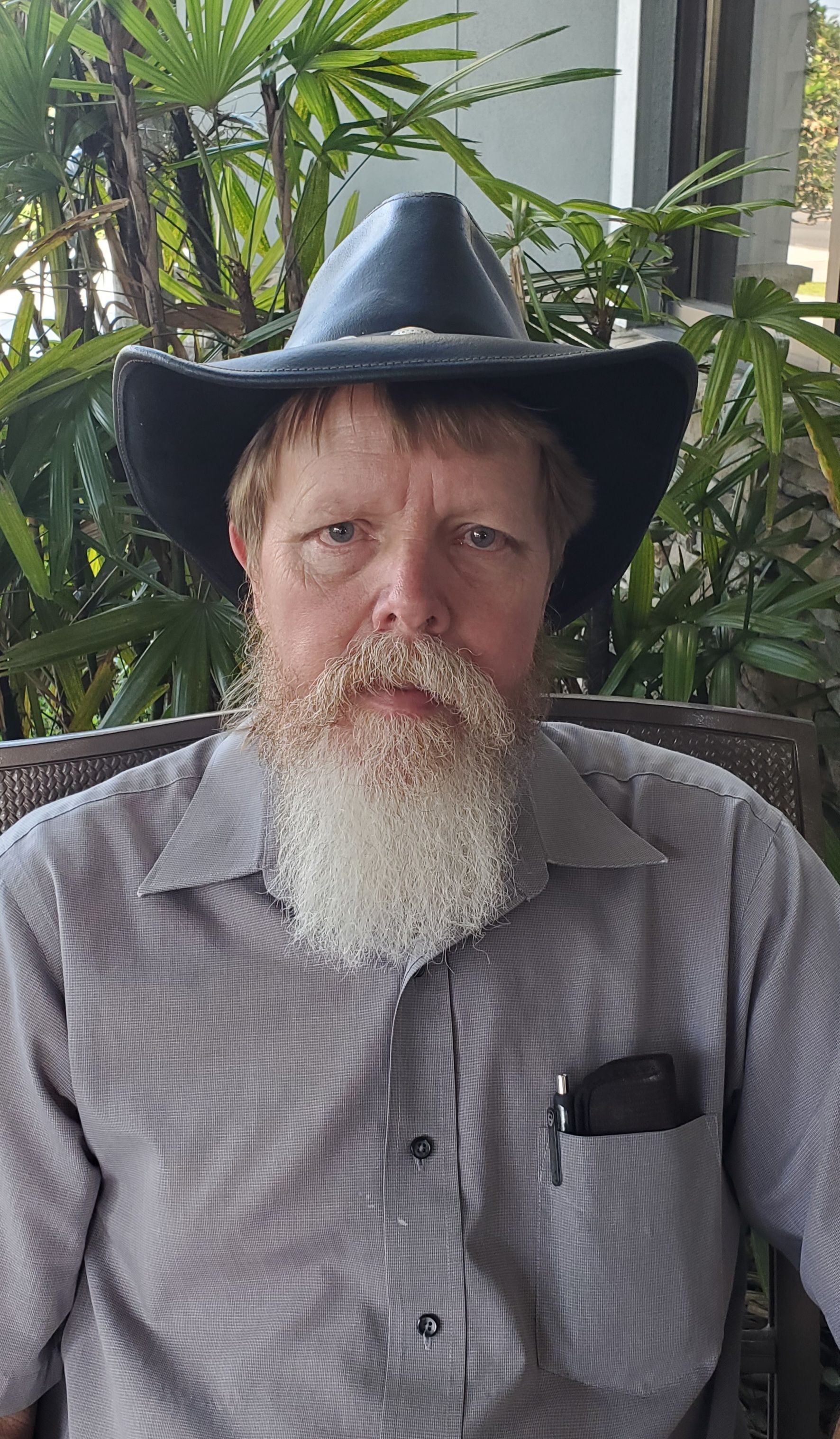 A man with a long, white beard and a cowboy hat sits outside, facing the camera. He wears a gray button-down shirt.