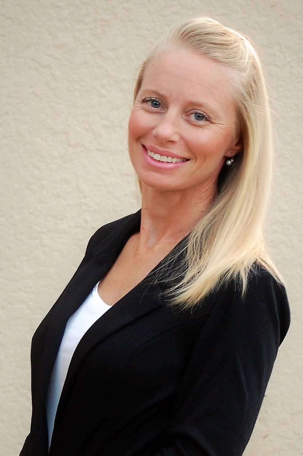 Blonde woman in a black blazer smiles at the camera, set against a beige wall.