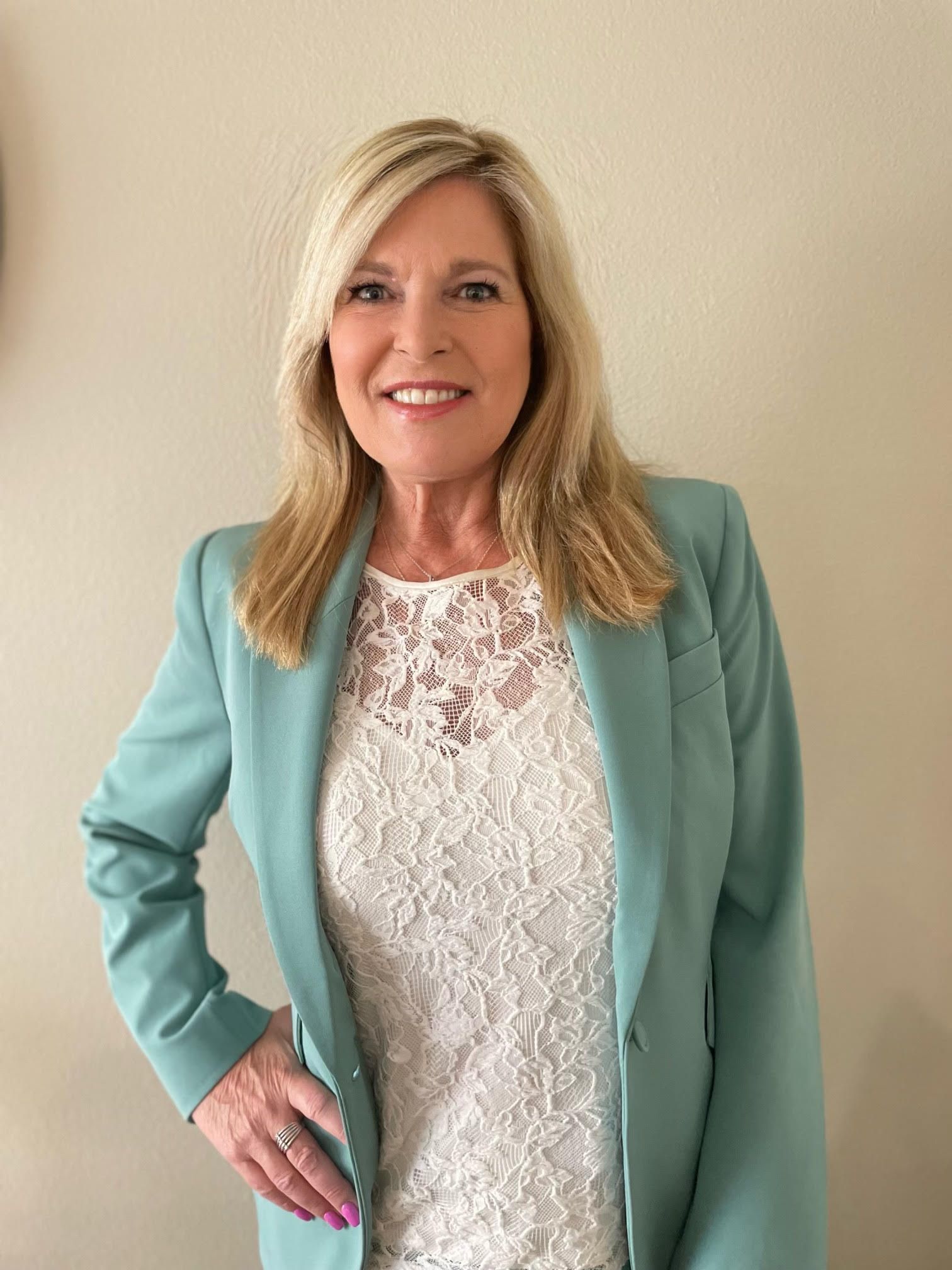 Woman with blonde hair wearing a teal blazer and a white lace top, smiling at the camera.