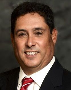 Man in a suit and tie, smiling. He has dark hair and appears to be in a professional setting.