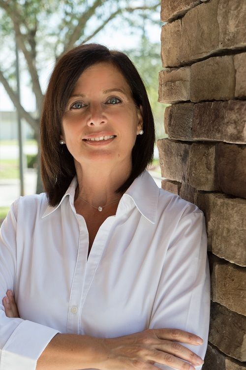Woman with dark hair smiles, arms crossed, leaning against a stone wall, wearing a white shirt outdoors.