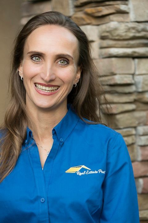 Woman with brown hair in a blue shirt with a logo smiles in front of a stone wall.