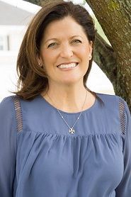 A woman wearing a blue shirt and a necklace is smiling.