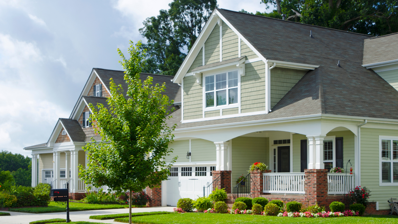Two-story houses with green siding and gabled roofs on a sunny day. A small tree is in front.