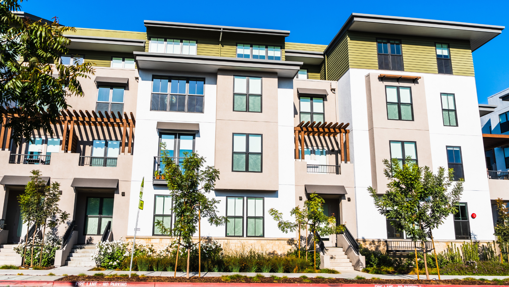 Multi-story modern townhouses with beige and white exteriors under a clear blue sky. Small trees and shrubs in front.