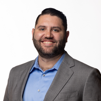 Man with beard smiles, wearing a blue shirt under a gray suit jacket against a white background.