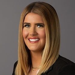 Woman with blonde hair smiles, wearing a black blazer against a grey background.