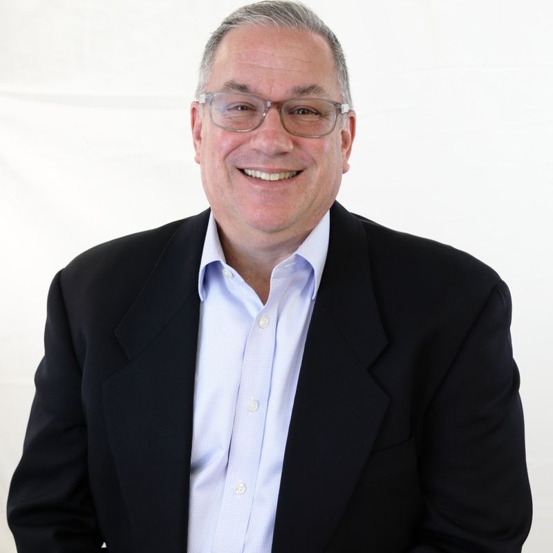 Man wearing glasses, black blazer, and blue shirt, smiles.