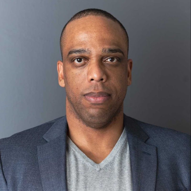 Man in blue blazer and gray v-neck shirt against a gray background, looking directly at the camera.