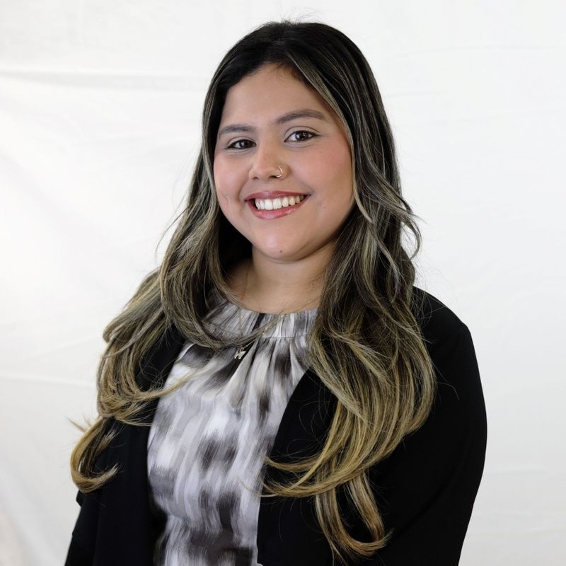 Woman smiling, long wavy hair, black blazer over a gray and white patterned top against white backdrop.
