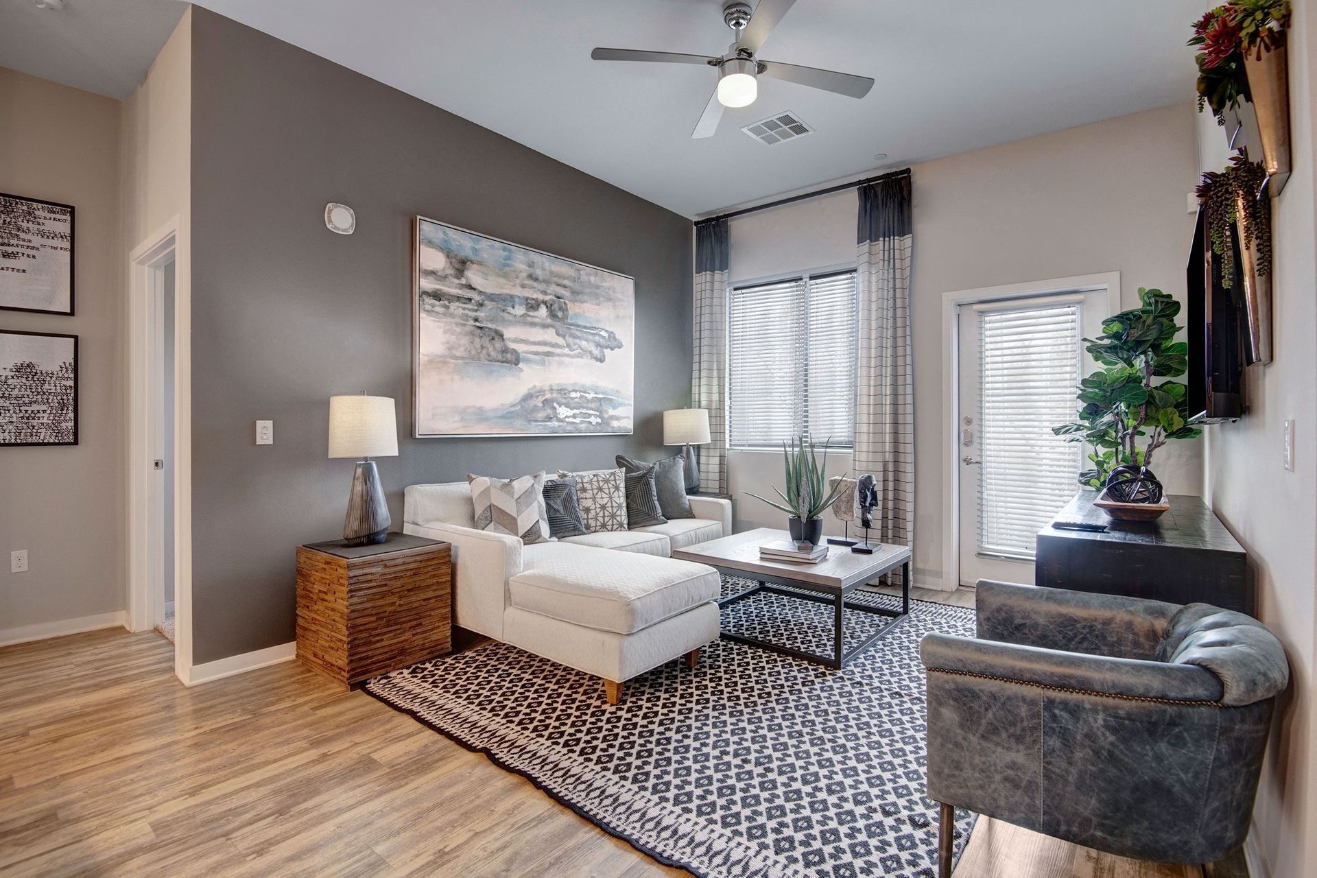 A modern living room here at Park on Central Apartments with a white sectional sofa, patterned rug, and gray accent chair. It features a large abstract painting, indoor plants, and two lamps, creating a cozy and elegant ambiance.