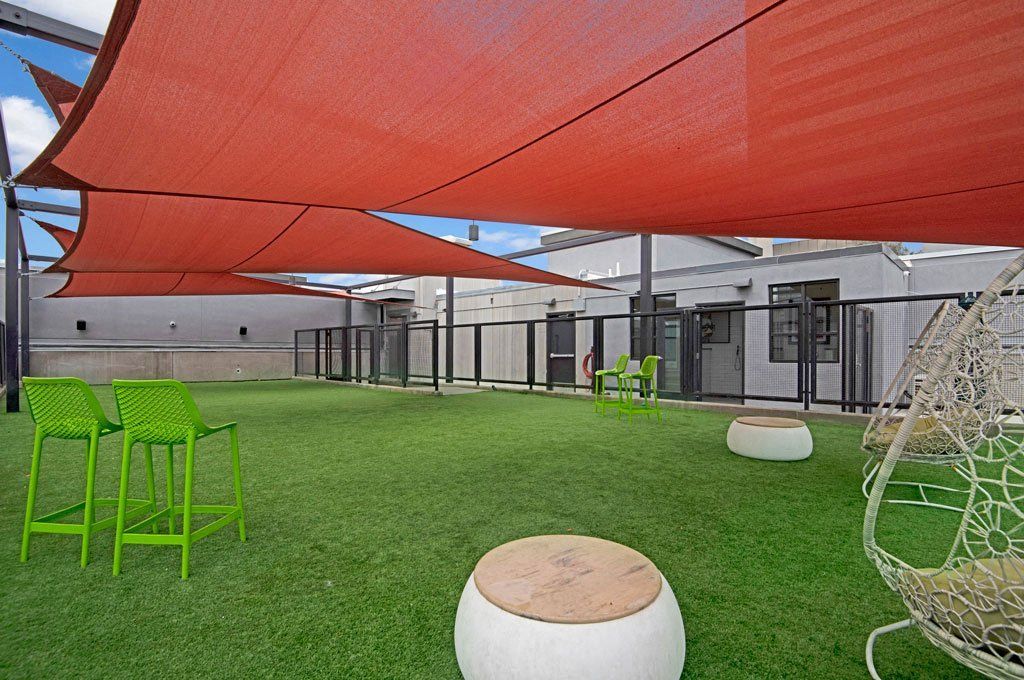 Spacious rooftop patio here at Park on Central Apartments with green artificial turf, red shade sails, modern green chairs, white round seats, and a wicker swing, creating a relaxing vibe.
