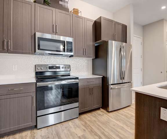 Modern kitchen here at Park on Central Apartments with dark wood cabinets, stainless steel appliances, and a sleek island with a sink. Warm wood flooring adds a cozy touch.