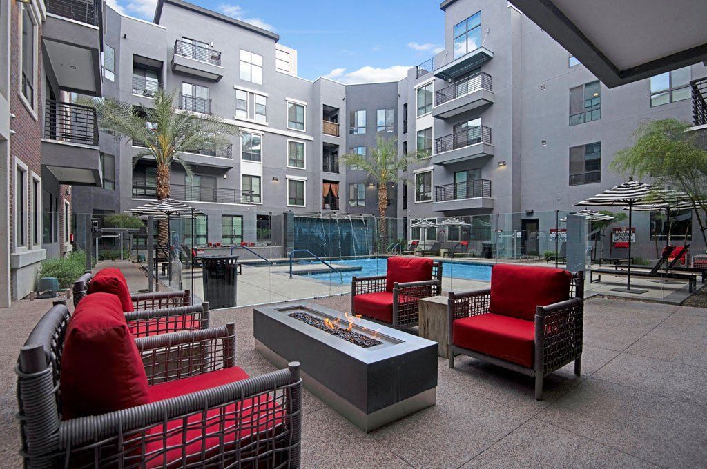 Modern Park on Central Apartment courtyard with a pool, surrounded by gray buildings. Red cushioned chairs and a fire pit create a cozy seating area, exuding a relaxed vibe.