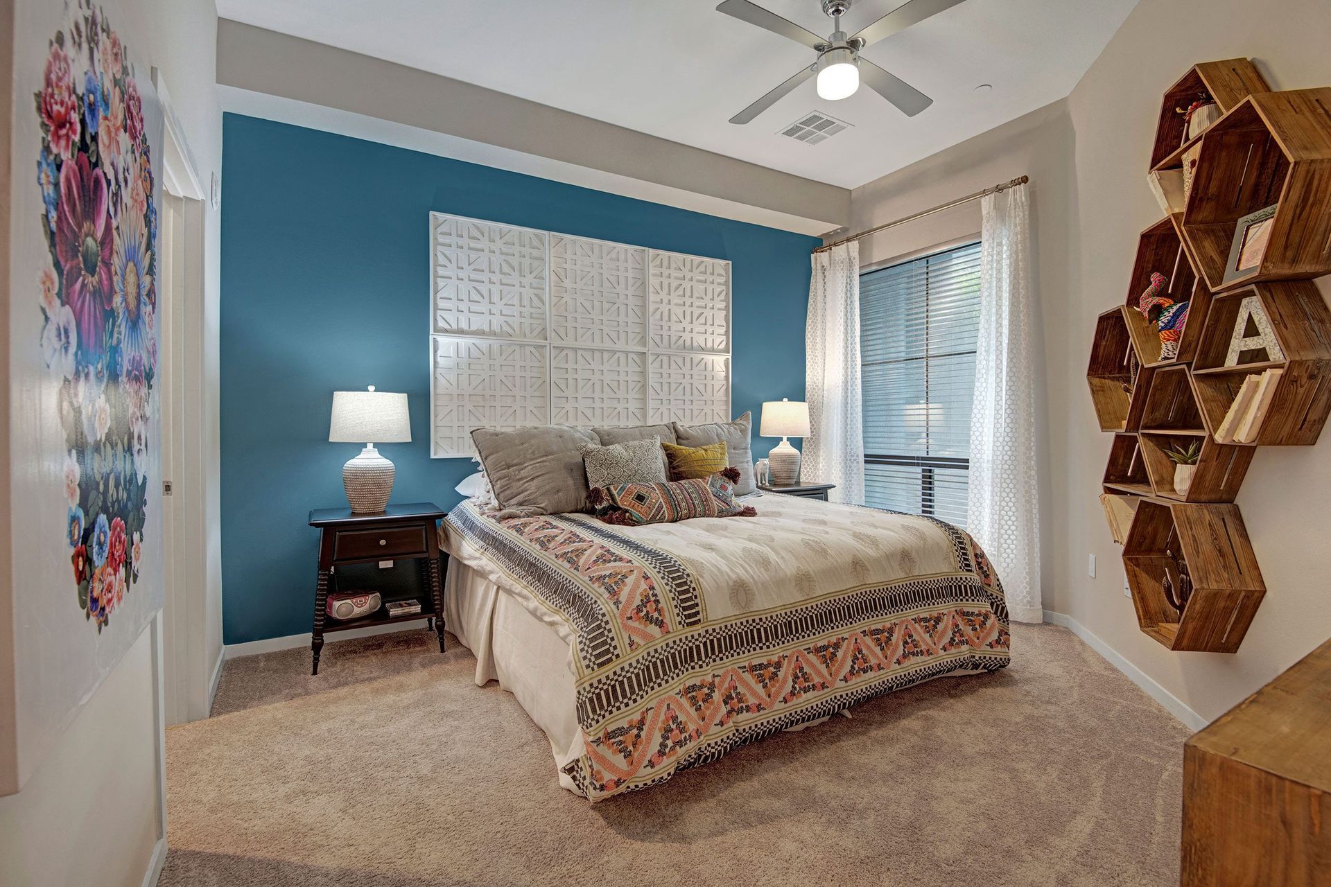 Cozy bedroom here at Park on Central Apartments with a bold blue accent wall, patterned bedding, and hexagonal wooden shelves. Warm lighting and floral wall art create a welcoming vibe.