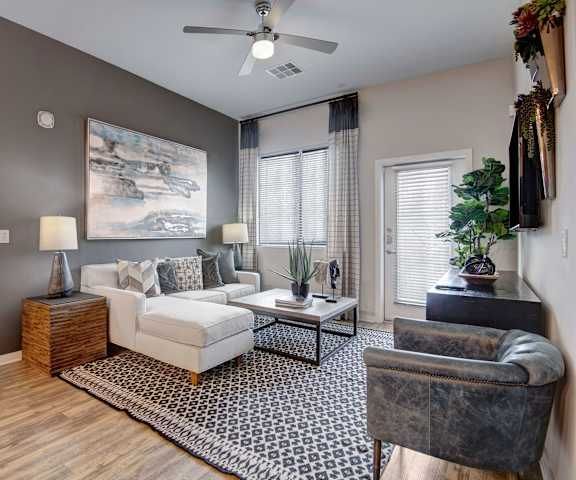 A modern living room here at Park on Central Apartments with a white sectional sofa, patterned rug, and gray accent chair. It features a large abstract painting, indoor plants, and two lamps, creating a cozy and elegant ambiance.