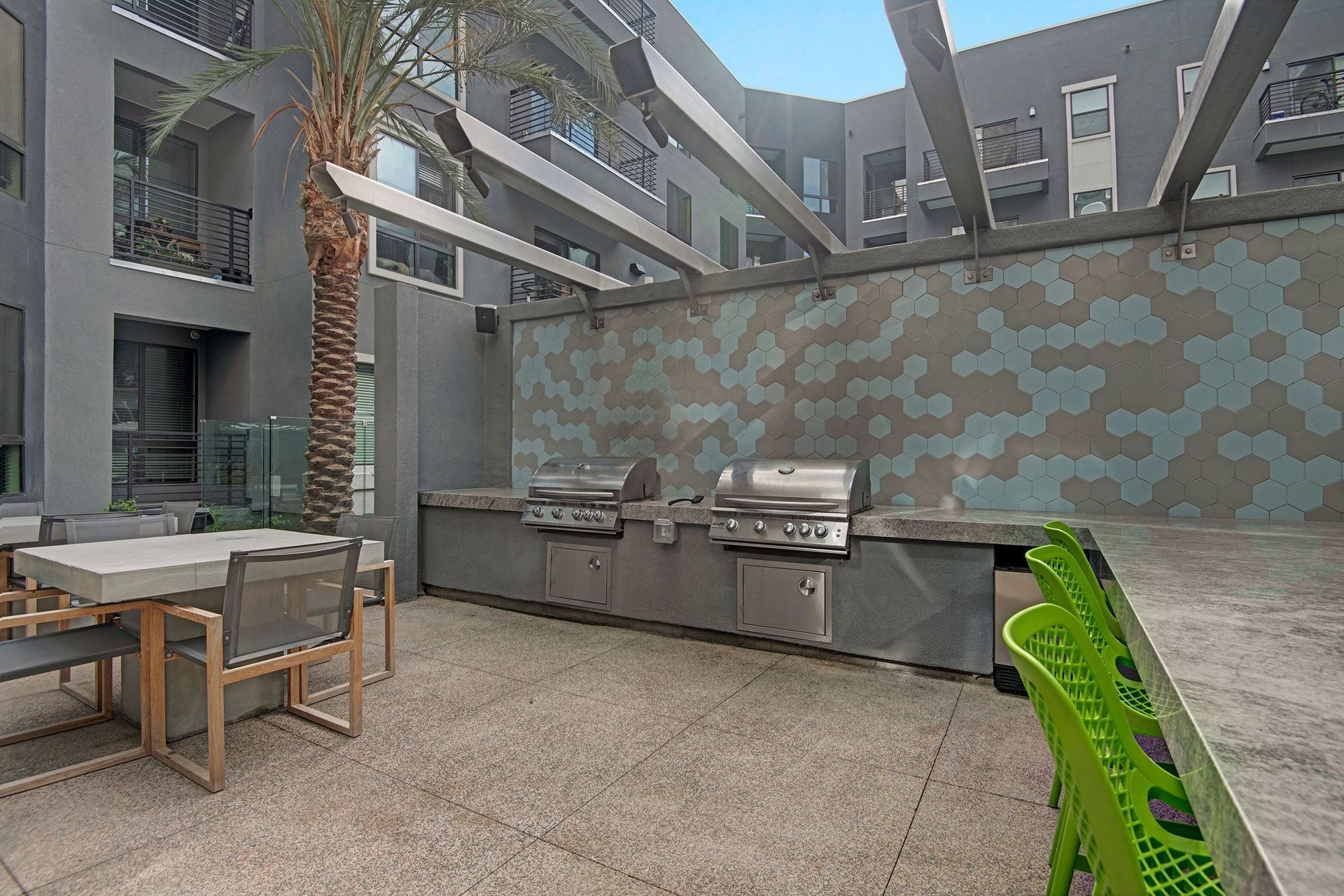 Modern outdoor courtyard here at Park on Central Apartments with sleek grills, a hexagonal tile wall, a dining area with gray chairs, and a vibrant mix of greenery and urban elements.