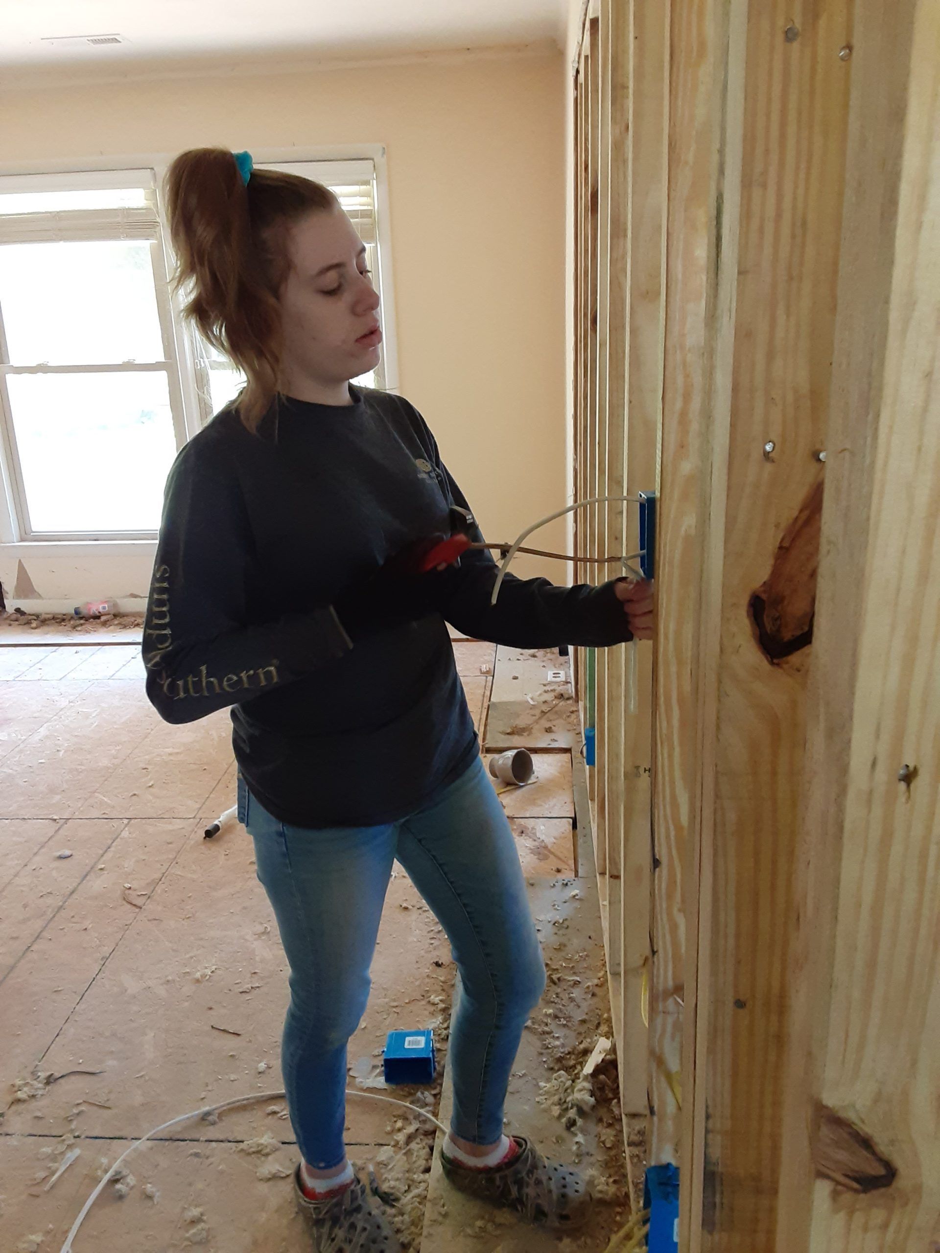 Woman wiring electrical outlet on wooden wall. Wearing jeans, long-sleeve shirt, and work gloves. Interior construction site.
