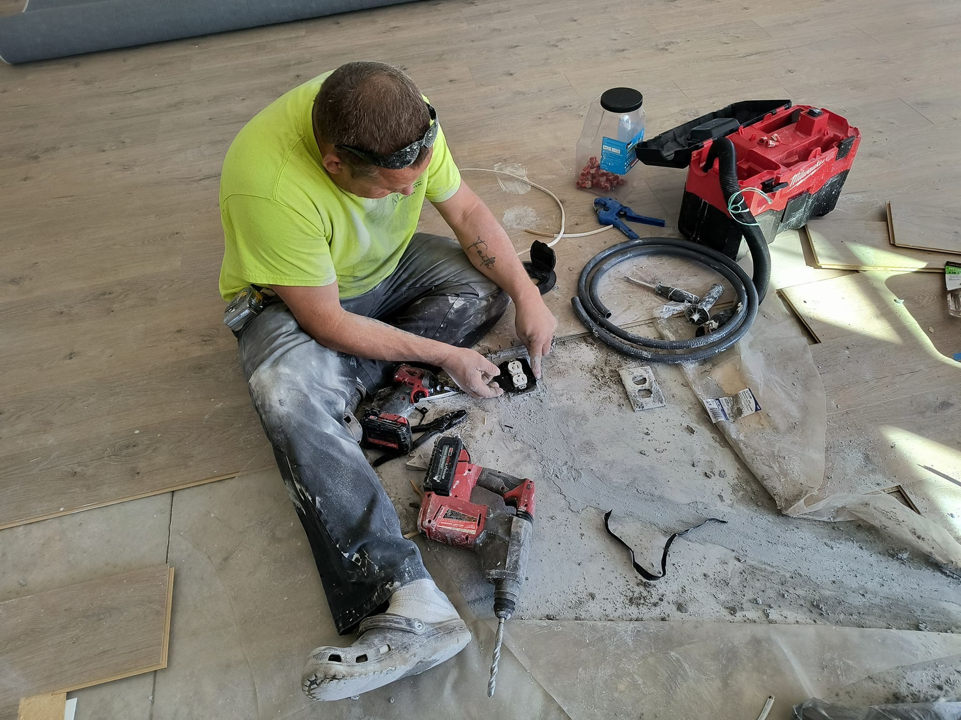 Person on floor surrounded by tools, working on a surface with debris. Light green shirt, jeans, safety glasses, and Crocs.
