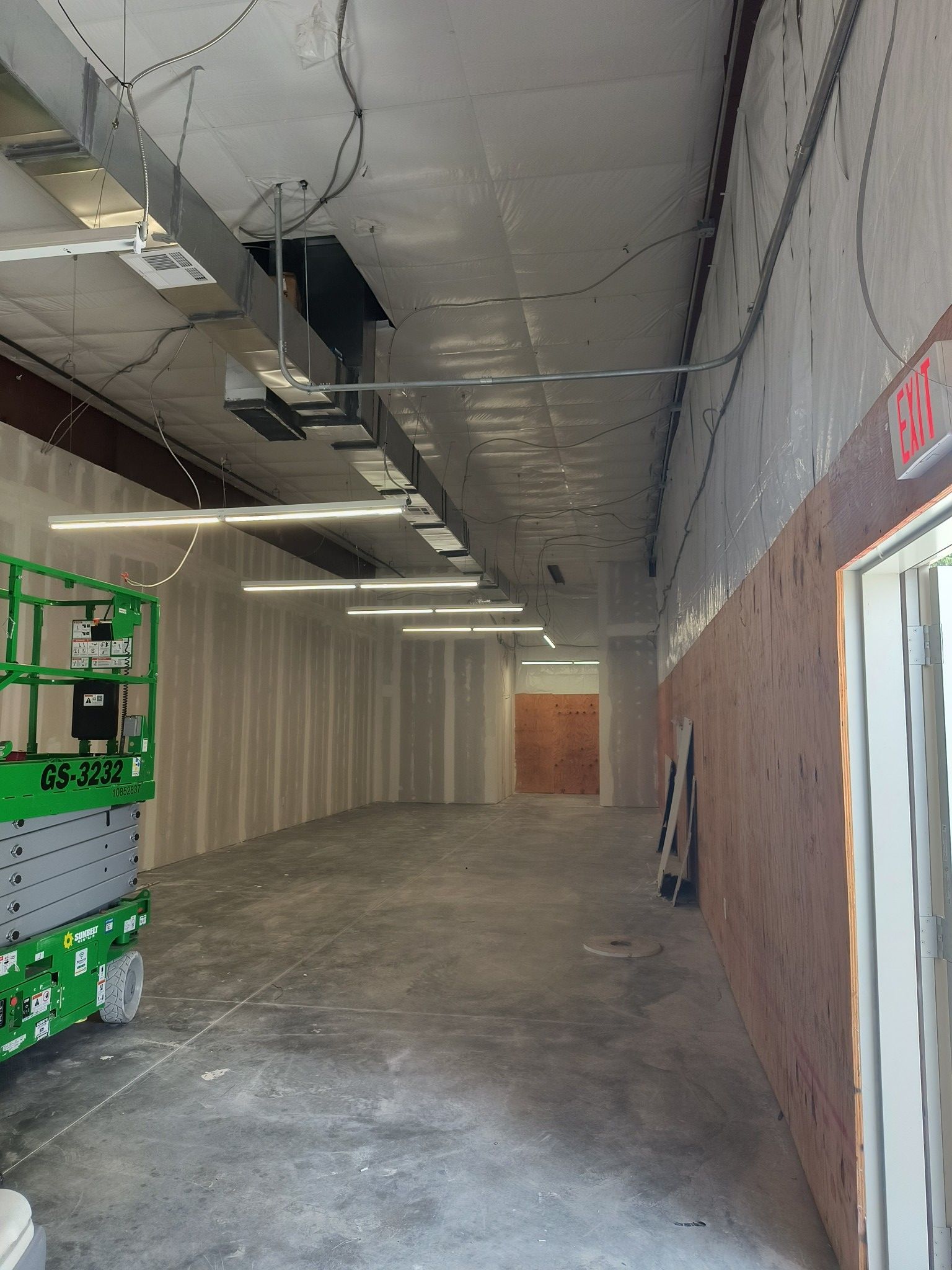 Interior view of a construction site. Concrete floor, drywall walls, overhead lights, and a scissor lift.