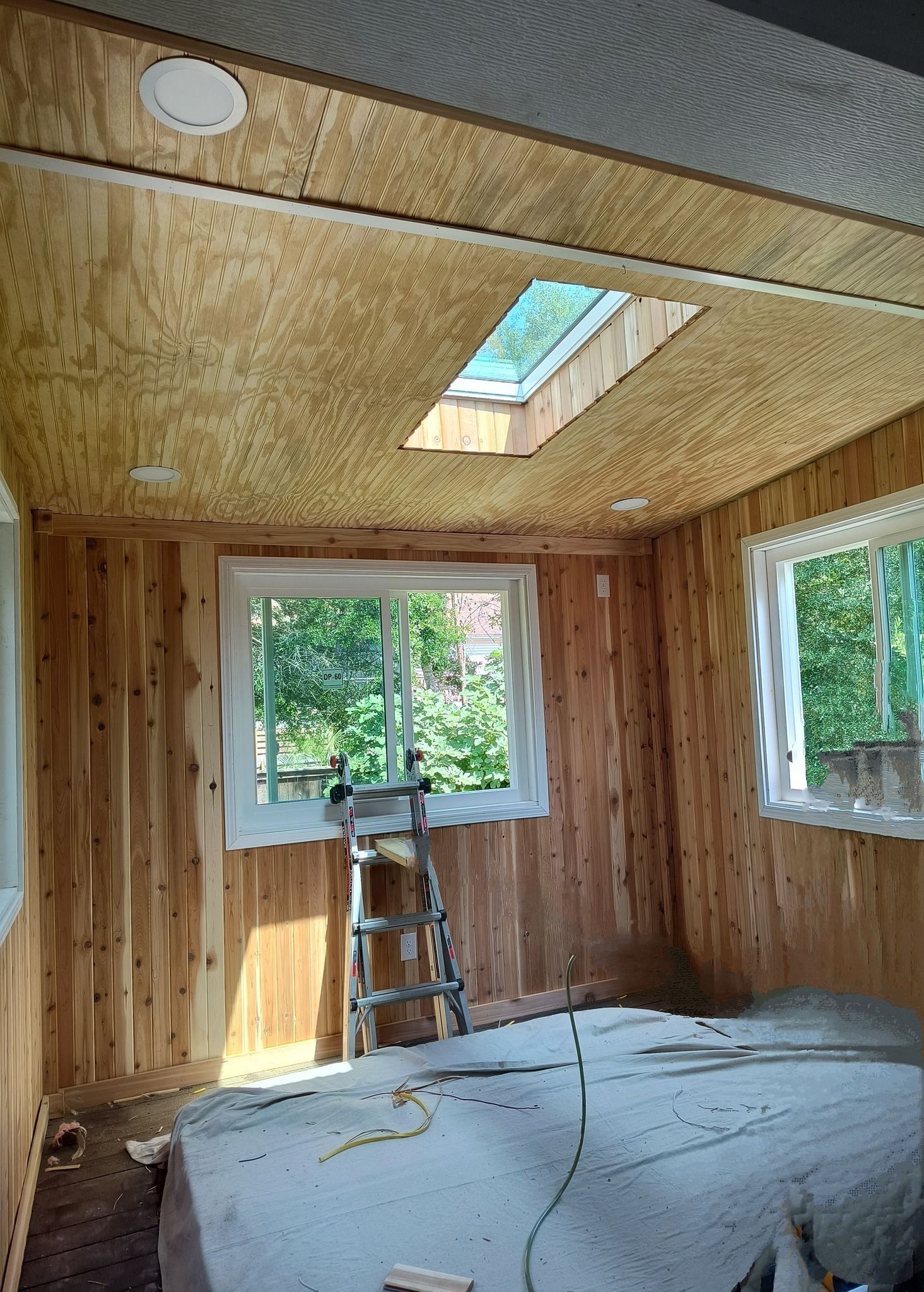 Room with wood paneling on walls and ceiling, a skylight, and windows. A ladder sits below the window.