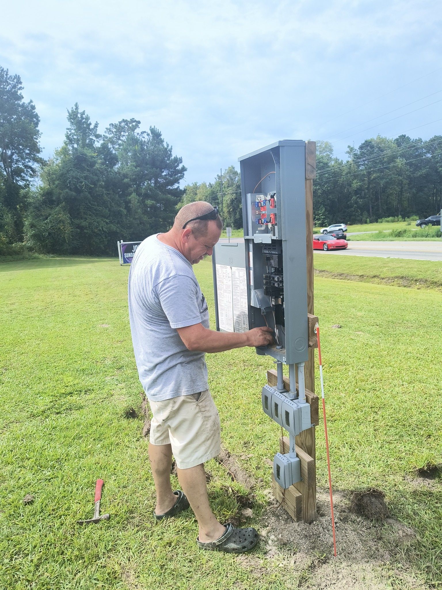 Man installing electrical panel on a wooden post in a grassy field.