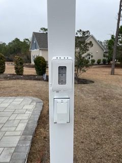 Outdoor utility pole with a mounted electrical outlet and white panel beside a paved driveway
