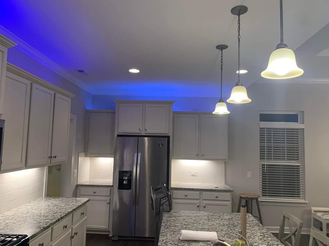A kitchen with white cabinets, stainless steel refrigerator, and pendant lights. Blue accent lighting on the ceiling.