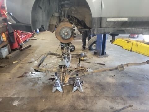 Car raised on a lift with front suspension components removed and scattered on the shop floor.