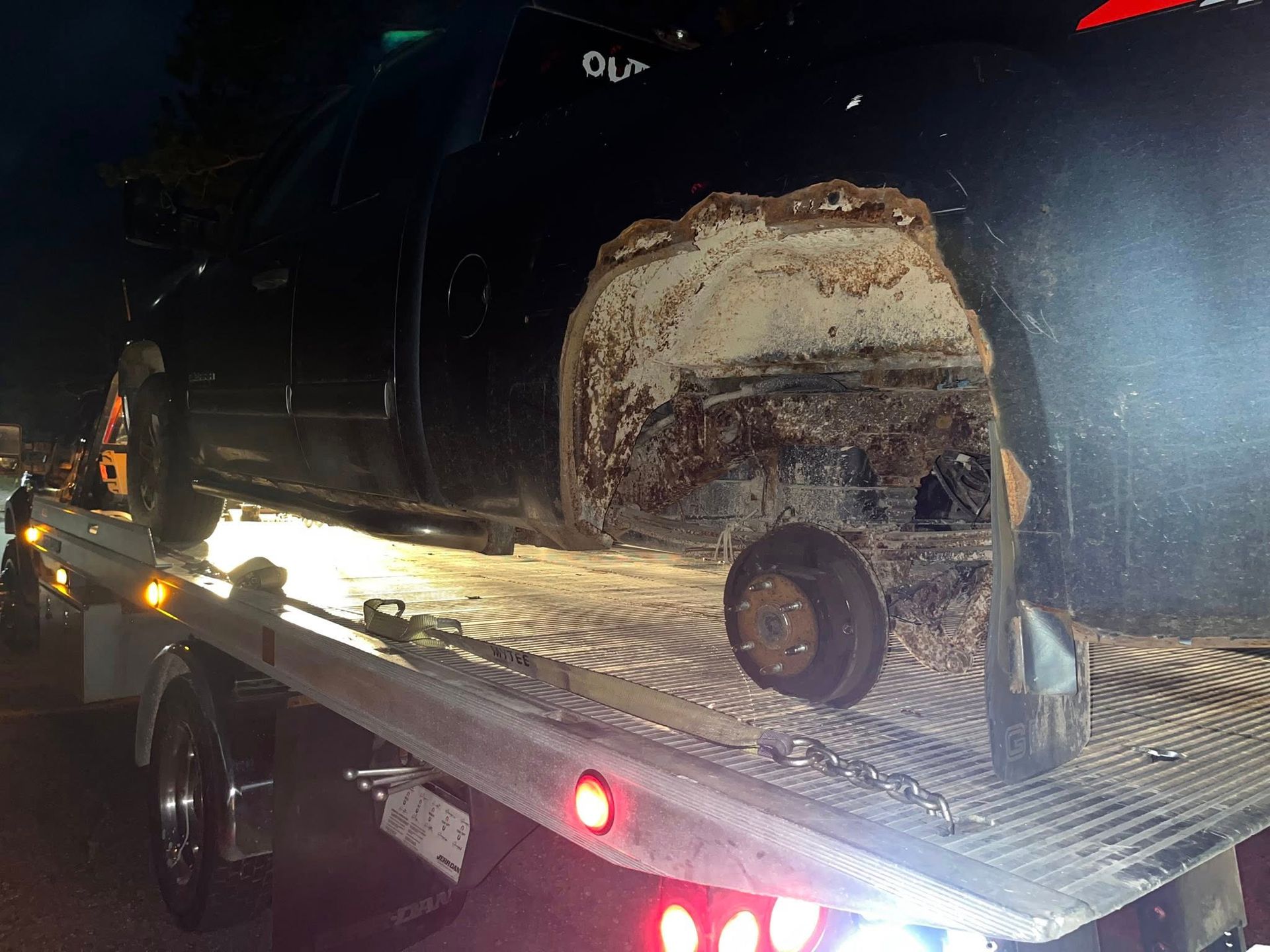 Black pickup truck on tow truck bed, significant rust visible around missing wheel.