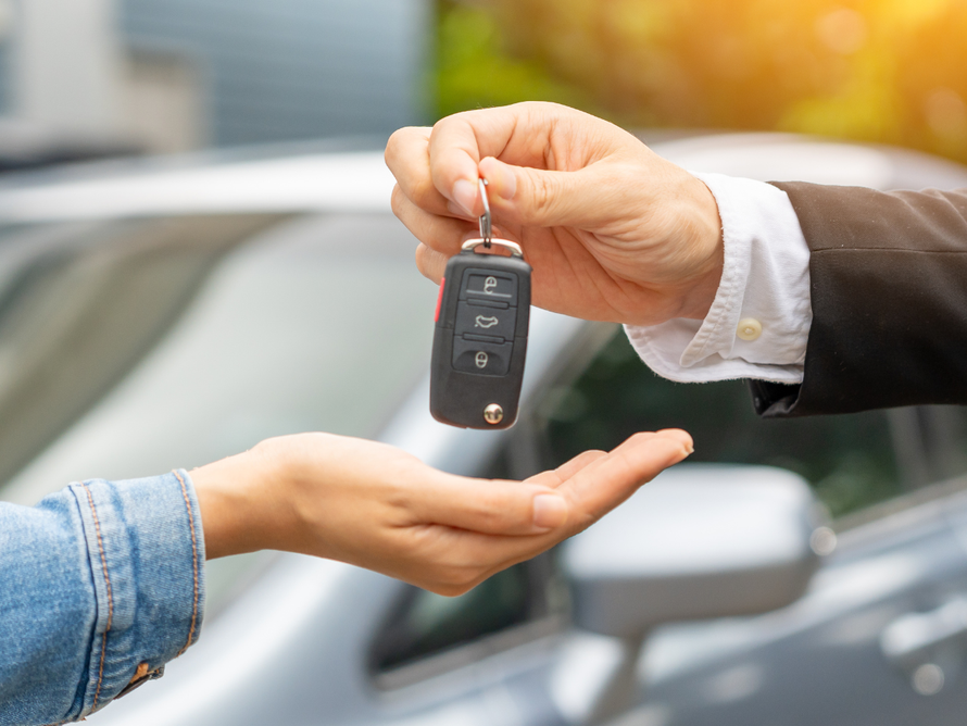 A hand in a business suit passes a car key fob to a waiting hand, with a blurred car in the background.