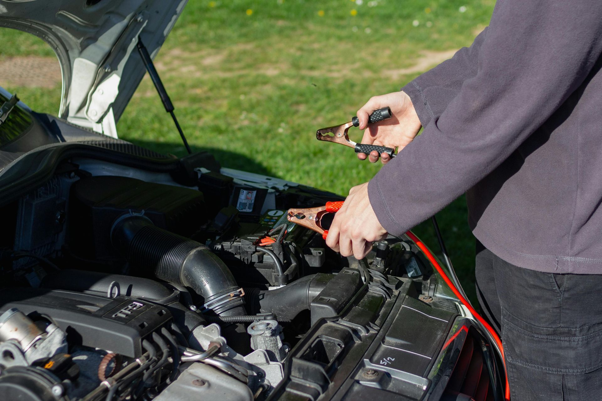 Person using jumper cables on a car battery under an open hood outdoors
