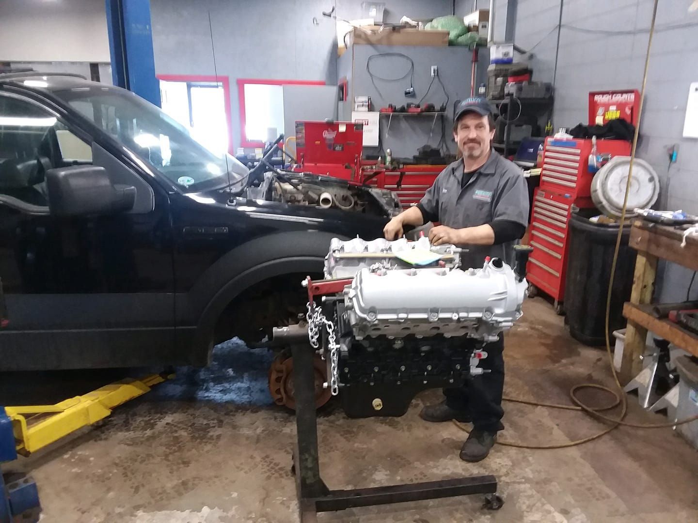 Mechanic stands next to a car and a rebuilt engine in an auto shop.