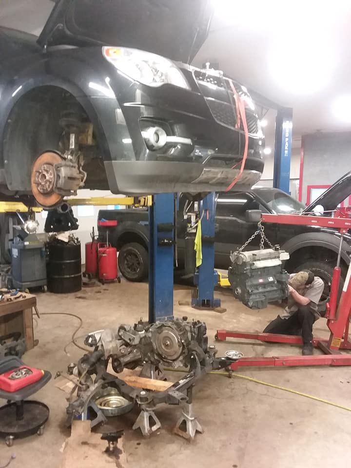 Car on a lift in a garage, engine removed. A mechanic works nearby.