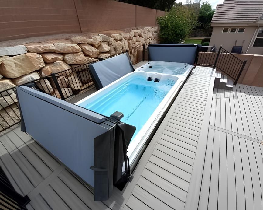 A long, rectangular swim spa with blue water is on a wooden deck. The cover is open.