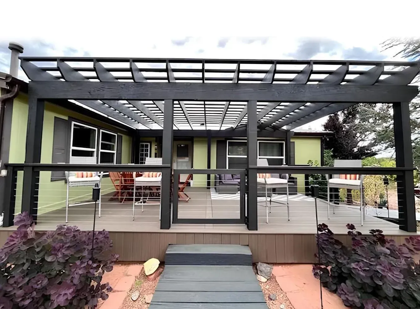 Green house with dark gray pergola over a deck, fronted by purple shrubs and a gray walkway.
