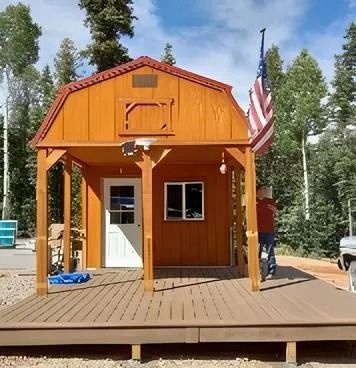 Wooden cabin with porch, US flag, person standing near. Orange exterior, blue sky, trees.