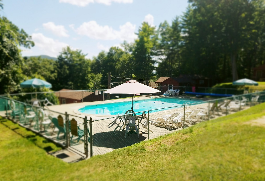 Outdoor swimming pool with blue water, white umbrellas, and lounge chairs beside a grassy lawn and trees