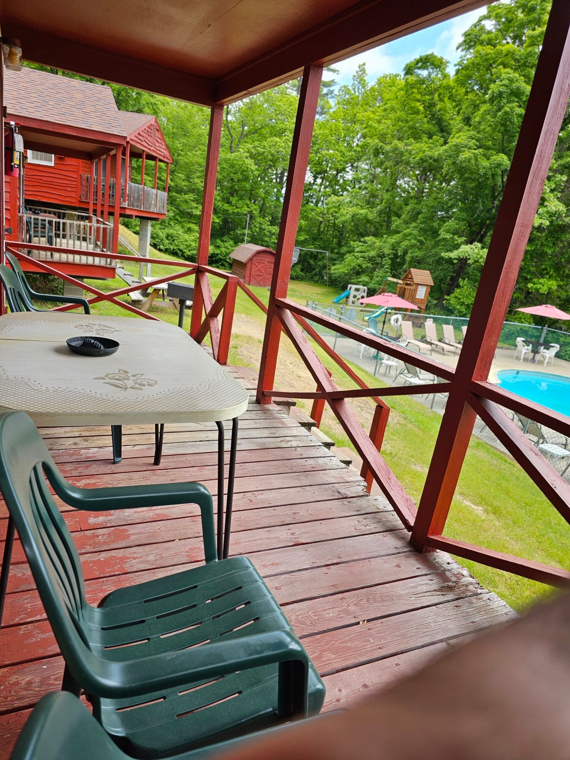 Porch overlooking a pool, with green chairs, a table, and a red cabin in a wooded setting