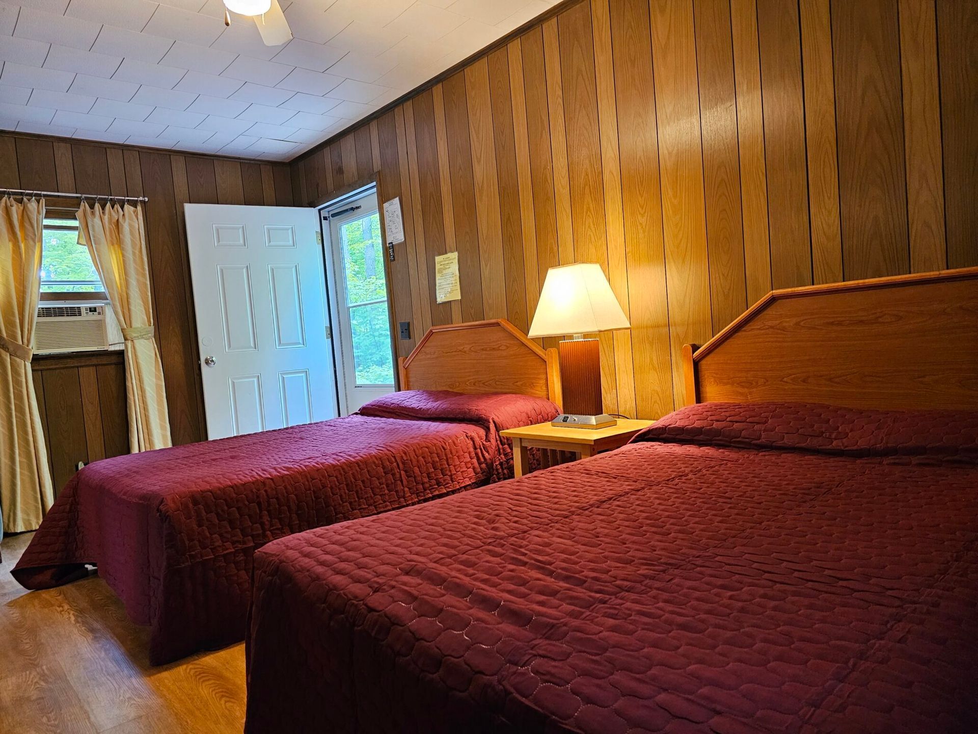 Wood-paneled bedroom with two red bedspreads, a lit lamp, and an open door to a bright hallway