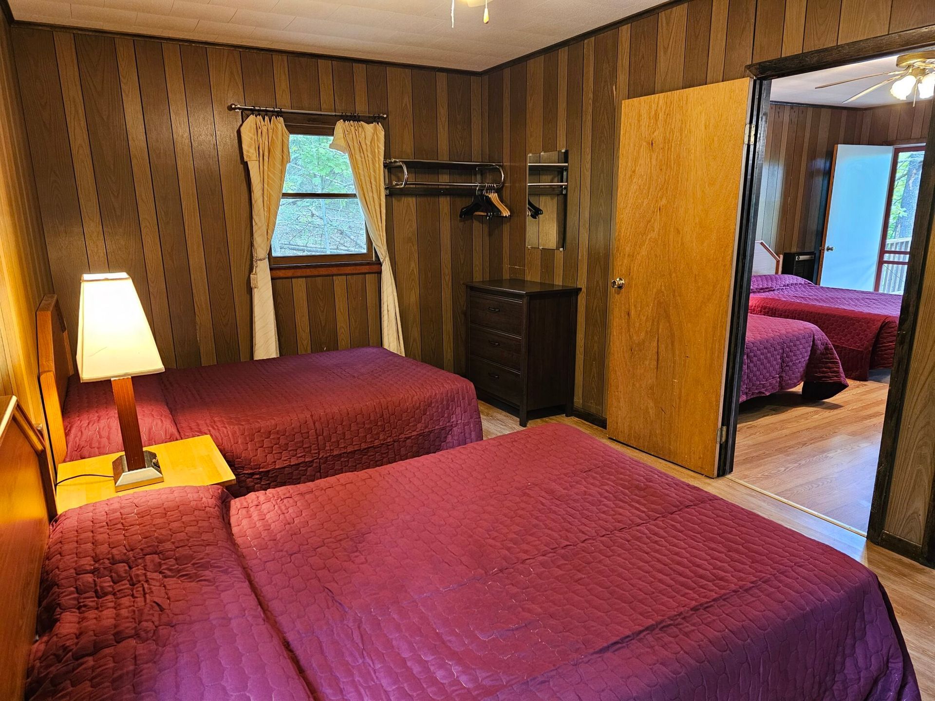 Cozy bedroom with two red beds, wood-paneled walls, lamp, closet, and adjoining room visible through an open door