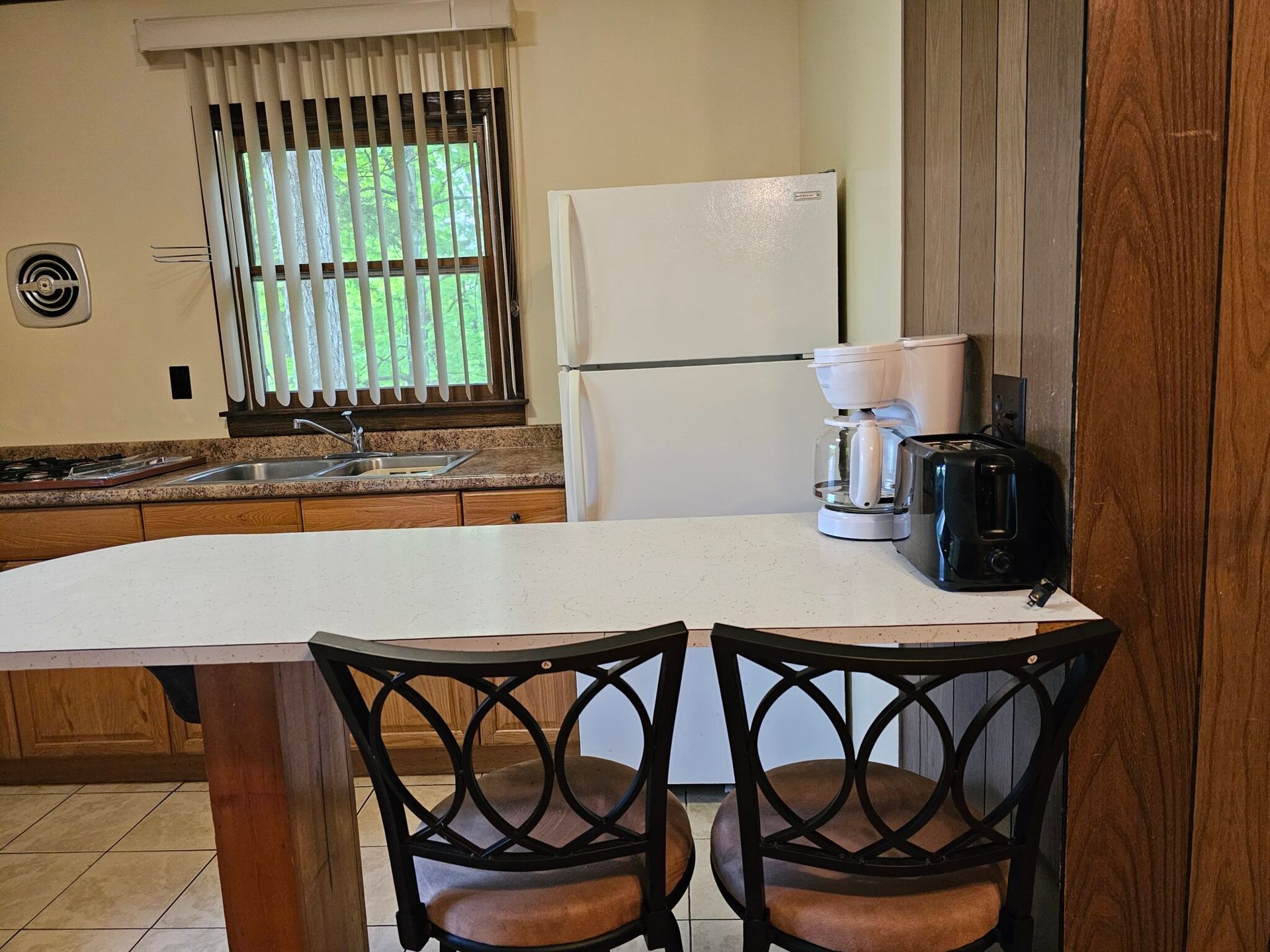 Small kitchen with white countertop, white fridge, coffee maker, and two chairs by a window.
