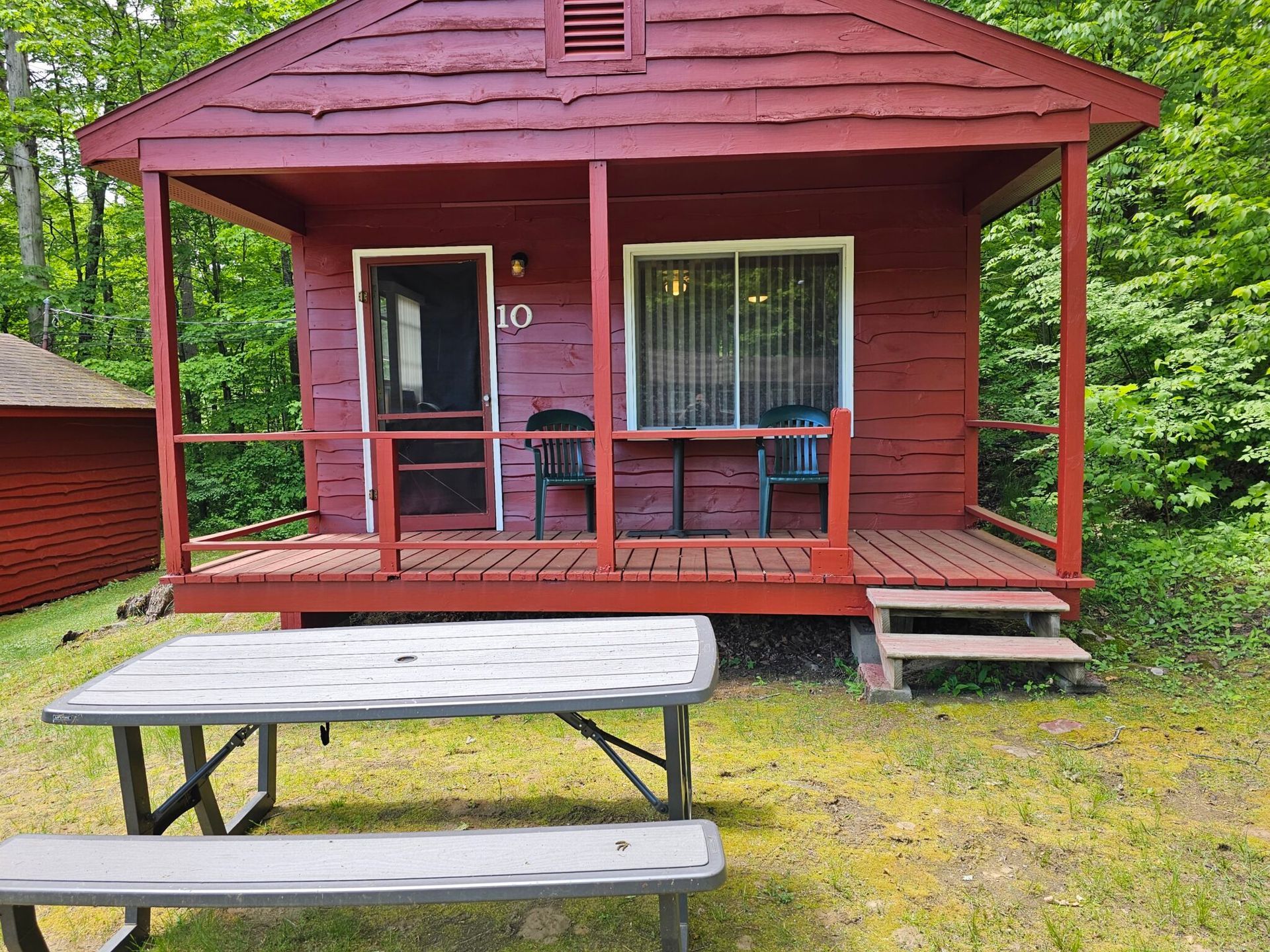 Red cabin porch with chairs, window, and picnic table in a grassy yard